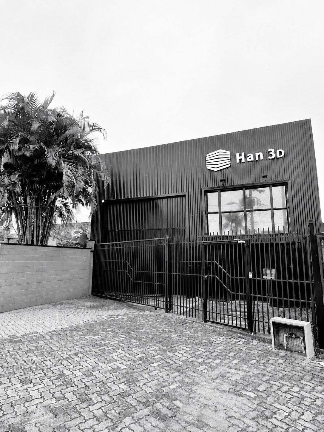 Fachada da sede industrial Han 3D em Novo Hamburgo, Rio Grande do Sul, fotografia em preto e branco, galpão moderno com fachada em ripas metálicas verticais escuras, logotipo Han 3D iluminado, portão de grade preto, palmeiras à esquerda, piso intertravado, céu claro, fotografia arquitetural realista